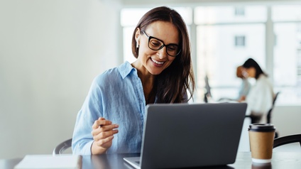 Nahaufnahme einer Person mit Brille, die vor einem aufgeklappten Laptop sitzt. Der Laptop steht auf einem dunkelbraunen Holztisch, daneben steht ein Coffee-to-go-Becher, ebenso liegen ein Notizblock und ein Smartphone auf dem Tisch