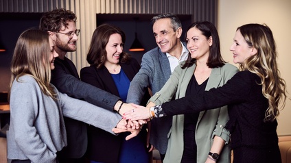 Eine Gruppe von Personen steht zusammen. Jede hat eine Hand ausgestreckt, die Personen führen die Hände in der Mitte zusammen und stapeln diese übereinander. 