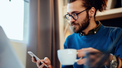 Bärtige Person mit Brille hält in einer Hand Tasse, in anderer Hand Smartphone, daneben aufgeklapptes Notebook