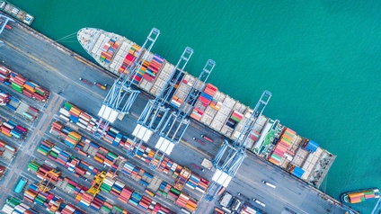 Vogelperspektive auf ein Containerfrachtschiff am Hafen mit unzähligen Containern, die mit Kränen auf das Schiff geladen werden.