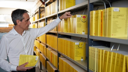Person mit weißem Hemd hält ein Rechtsbuch in Händen und greift mit der anderen Hand zu einem weiteren Buch in einem Regal einer Bibliothek