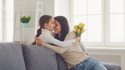 Kind mit gelbem Tulpenstrauß in Hände umarmt erwachsene Person auf Sofa sitzend innig