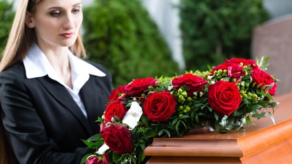 Eine Bestatterin steht vor einem Holzsarg und rückt den Kranz aus roten Rosen zurecht