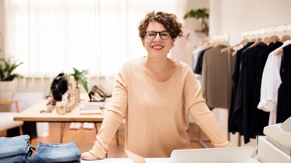 Lächelnde Person mit Brillen steht an Tisch mit aufgeklapptem Laptop, Zahlungsgerät und Stapeln von Jeans, im Hintergrund Kleiderstangen und Tisch mit modischen Accessoires