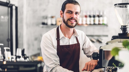 Person mit Bart und geschlossenen Haaren steht mit Arbeitskleidung  vor einer Kaffeemaschine und bereitet Kaffee zu