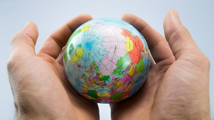 Nahaufnahme von zwei Händen, in denen sie einen kleinen Globus hält. Der Hintergrund ist grau, auf den Globus fällt Licht. 