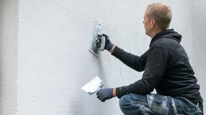 Rückenansicht einer Person in Arbeitskleidung und mit Handschuhen, die mit einer Spachtel eine Fassade eines Gebäudes verputzt