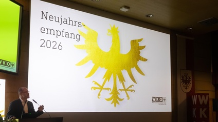 Eine Person steht am Rednerpult und spricht in ein Mikrofon. Hinter der Person ist eine große Leinwand mit der Aufschrift „Neujahrsempfang 2026“ und einem großen goldenen Adler zu sehen. Im Raum sitzen und stehen mehrere Personen an Stehtischen und hören zu. Der Raum ist hell beleuchtet und als Veranstaltungssaal erkennbar.
