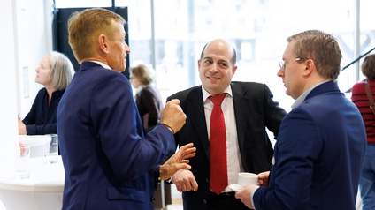 3 Männer in Anzügen im Gespräch an einem Stehtisch