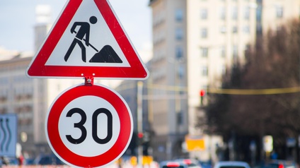Zwei Straßenschilder im Fokus: Ein Baustellen- und ein 30-er-Zonene-Schild, im Hintergrund verschwommen Autos in Rückansicht.