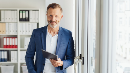 Mann mit blauem Sakko und Dreitagebart lehnt an einer Fensterfront und hat ein Tablett in der Hand, im Hintergrund sieht man ein Regal mit Ordnermappen