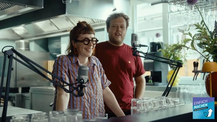 Frontale Aufnahme von zwei Personen, die hinter einem Tresen stehen. Vor den Personen sind zwei Mikrofone angebracht. Hinter ihnen ist eine Küche