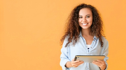 Eine Person mit lockigem, langem dunkelbraunem Haar steht vor gelbem Hintergrund. Sie hält mit beiden Händen ein Tablet. Die Person ist im rechten Bildrand.