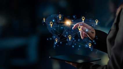 In der Mitte des Bildes ist eine Grafik der Kontinente der Welt. An manchen Stellen sind Icons für Lokalisierungen, die miteinander verbunden sind. Die rechte Hand einer Person zeigt mit einem Stift auf eine Stelle