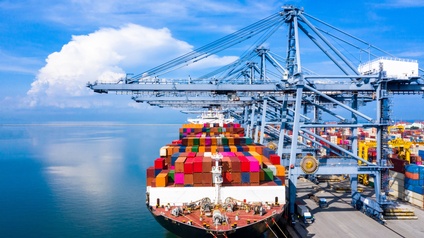 Ein Frachtschiff mit gestapelten Containern ist an einem Hafen angedockt. Direkt neben dem Schiff sind Hafenkräne. Im Hintergrund sind viele weitere Container