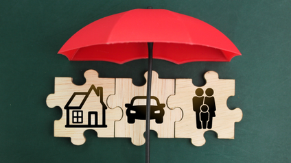 Roter Regenschirm vor 3 Holzpuzzleteilen mit Haus-, Auto- und Menschensymbolen