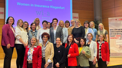Mehrere Frauen stehen zum Gruppenfoto auf und vor einer Bühne. Im Hintergrund kann man auf einer Leinwand den Schriftzug Women in Insurance Klagenfurt lesen.