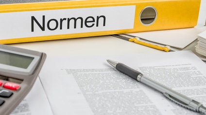 Gelber Ordner mit Beschriftung Normen auf Längsseite liegend, davor Stift auf Dokument und Ausschnitt eines Taschenrechners