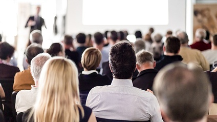 Mehrere sitzende Personen in Rückenansicht, im Hintergrund verschwommen Person auf Bühne