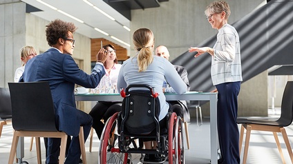 Hier sieht man eine Meetingsituation mit einer Rollstuhlfahrerin und anderen vielfältigen Personen.