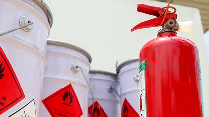 Im Vordergrund roter Feuerlöscher, im Hintergrund mehrere Fässer mit Gefahrengutsticker