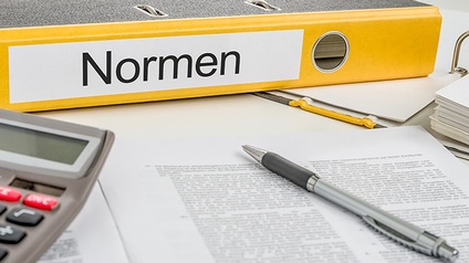 Gelber Ordner mit Beschriftung Normen auf Längsseite liegend, davor Stift auf Dokument und Ausschnitt eines Taschenrechners