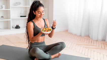 Junge Frau sitzt auf einer Yogamatte und isst einen Salat.
