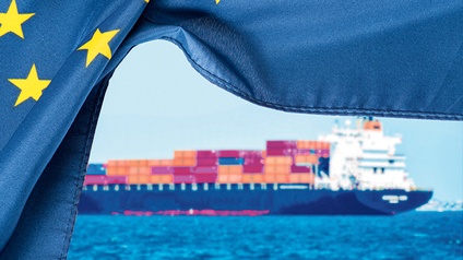 Man sieht eine EU-Fahne (blau, links drei gelbe Sterne) und dahinter ein großes Frachtschiff im Meer.