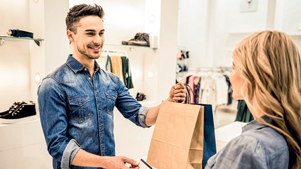 Ein junger Mann steht in einem Kleidungs/Schuh-Geschäft und reicht einer jungen Frau ein braunes Einkaufssackerl (Verkaufssituation).