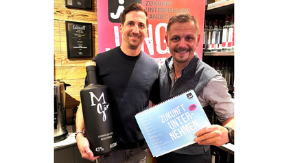Zwei Personen stehen nebeneinander in einem Geschäft oder Showroom mit Holzregalen und Flaschen im Hintergrund. Beide lächeln in die Kamera. Der Mann links hält eine große schwarze Flasche mit der Aufschrift „M Gin“ in den Händen. Der Mann rechts hält einen blauen Block oder ein Heft mit der Aufschrift „Zukunft Unternehmen“. Im Hintergrund sind Regale mit Flaschen sowie Schilder und Auszeichnungen an der Wand zu sehen. Die Szene wirkt wie eine Präsentation oder ein gemeinsamer Termin im geschäftlichen Umfeld.