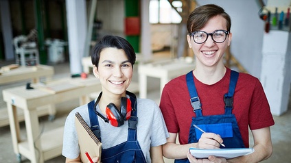 Zwei lächelnde Personen in blauen Arbeitslatzhosen mit Stiften und Notizblöcken in Tischlerwerkstatt stehend