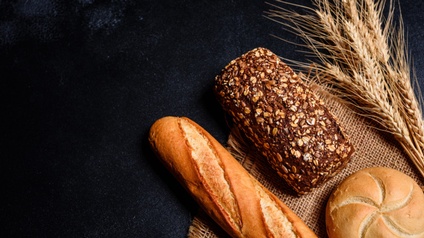 Backwaren wie Baguette, Brot und Semmel gemeinsam mit Stoff und Weizenähre auf einem dunklen Untergrund platziert, Vogelperspektive