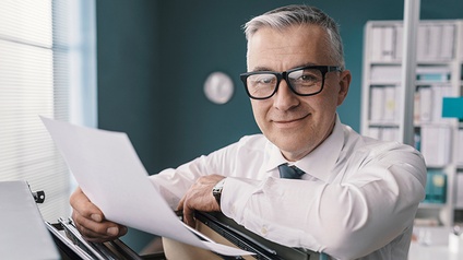 Sanft lächelnde Person mit Brille lehnt über Aktenlade und hält Dokument in einer Hand