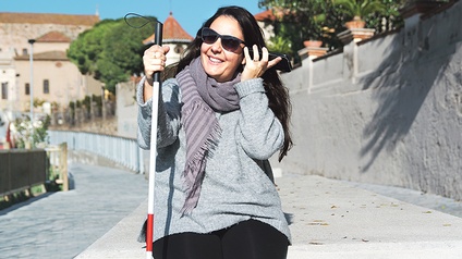 Frau mit Blindenstock, Sonnenbrille und Smartphone am Ohr  lacht