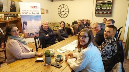 An einem langen Tisch in einem Lokal sitzen sich mehrere Personen gegenüber. Alle Personen blicken in die Kamera. Im Hintergrund steht ein Werbebanner, auf dem WKO im Bezirk steht sowie eine Aufnahme der Stadt Wien ist