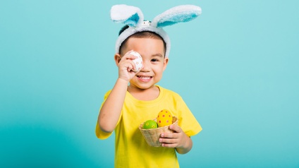 Halbporträt eines Kindes, das einen kleinen Korb in der linken Hand hält. Im Korb sind bemalte Eier. Mit der rechten Hand hält es sich ein bemaltes Ei vor das rechte Auge. Auf dem Kopf trägt es Plüschhasenohren