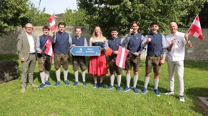 Neun Personen stehen auf einer Grünfläche vor Bäumen und einer Mauer. In der Bildmitte halten zwei Personen ein Schild mit der Aufschrift „Team AUSTRIA“. Auf dem Schild sind zusätzlich die Logos „WorldSkills Shanghai 2026“ und „SkillsAustria“