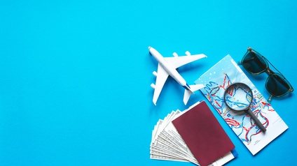Top-Shot auf Reiseaccessoires und -gegenstände auf blauem Hintergrund: Ein Modell eines Flugzeuges, eine Sonnenbrille, ein Reisepass mit Dollarscheinen sowie Flugticket und ein Stadtplan mit Lupe.