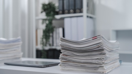 Auf einem weißen Tisch ist ein Dokumentenstapel. Daneben sind ein Tablet und ein weiterer Dokumentenstapel. Im Hintergrund ist ein Regal mit Ordnern