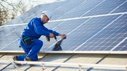 Person in blauem Arbeitsoverall mit weißem Helm kniet mit Bohrmaschine in Hand vor Solarpanelen und befestigt diese
