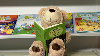 Ein hellbrauner, kuscheliger Teddybär sitzt auf einem Bücherregal und hält in den Händen ein Kinderbuch. Hinter ihm sind in einem Regal weitere Kinderbücher zu sehen.