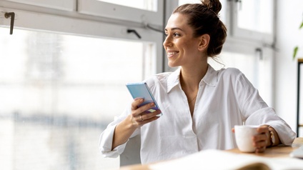 Lächelnde Person blickt aus Fenster an Tisch sitzend mit Smartphone in der Hand