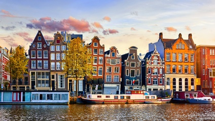 Aufnahme verschiedener, bunter, mehrstöckiger Häuser nebeneinander in Amsterdam. Vor den Häusern fließt ein Gewässer. Auf dem Wasser sind Boote