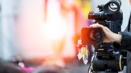 Fokus auf eine Filmkamera, die von einer Person betrieben wird, dahinter zeigt sich ein buntes Bokeh in der Unschärfe