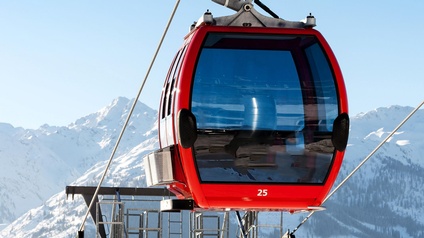 Eine rote Gondel mit der Nummer 25 an Seil hängend unter blauem Himmel, im Hintergrund verschneite Berglandschaft