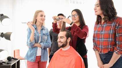 Eine lächelnde Person mit braunen Haaren, Bart und Umhang sitzt. Um die Person stehen vier Personen. Eine der Personen hält eine Haarsträhne der sitzenden Person und schneidet mit einer Schere die Spitzen. Die anderen Personen sehen zu