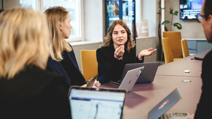 Drei Frauen und ein Mann sitzen mit aufgeklappten Laptops an einem Tisch und unterhalten sich