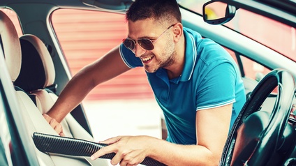 Person mit kurzen Haaren, Bart und Sonnenbrille bückt sich über einen Fahrersitz eines Fahrzeuges und hält dabei freudig einen Staubsauger
