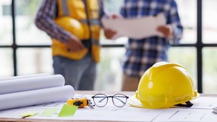 Im Vordergrund liegt ein Bauplan auf einem Tisch. Auf dem Plan liegen Papierrollen, eine Brille, ein Bauhelm und ein Maßband. Im Hintergrund stehen zwei Personen nebeneinander. Die linke hält einen Plan