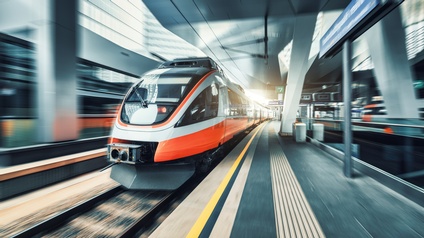 Auf Gleisen fährt ein einstöckiger, rot-grauer Zug. Rechts neben den Gleisen ist ein Bahnsteig